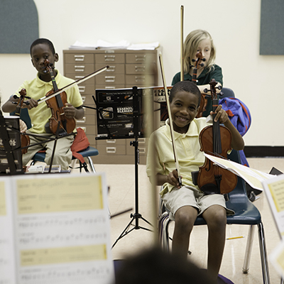 A child smiles while holding up a violin meanwhile others are practicing in the background