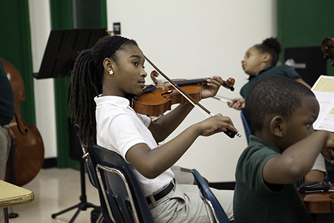 A woman with braided hair in a white polo shirt looks off to her right as she plays the violin
