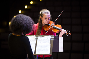 A woman with long blonde hair and red shirt looks down as she plays the violin