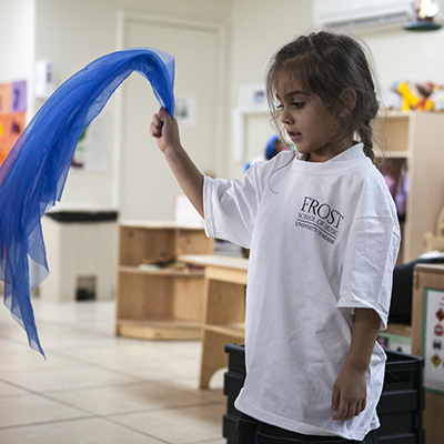 A young girl in a white Frost School of Music shirt waves around a piece of blue fabric